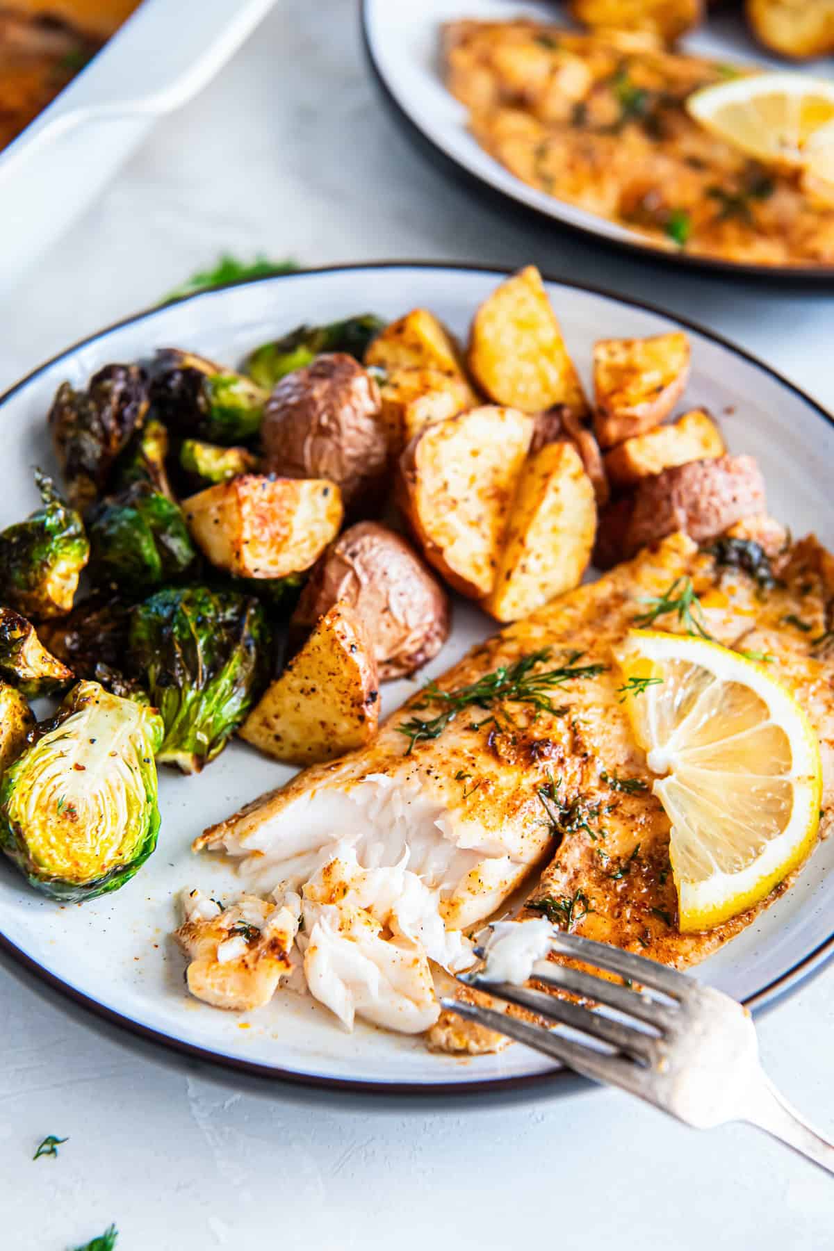 Plated easy baked fish with lemon and dill, served with roasted baby potatoes and Brussels sprouts, with a fork flaking the tender fish.