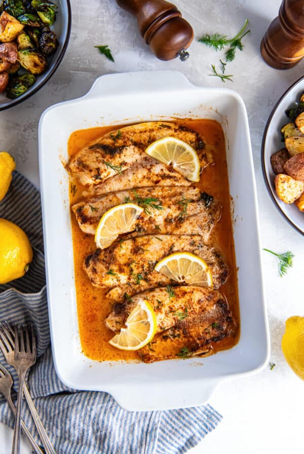 Easy baked fish fillets in a white baking dish with lemon slices and dill, surrounded by side dishes, whole lemons, and salt and pepper shakers.