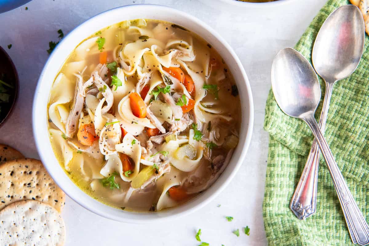 A bowl of homemade chicken noodle soup with shredded chicken, wide egg noodles, carrots, and celery in a golden broth, served with crackers, two spoons, and a green napkin on a light surface.