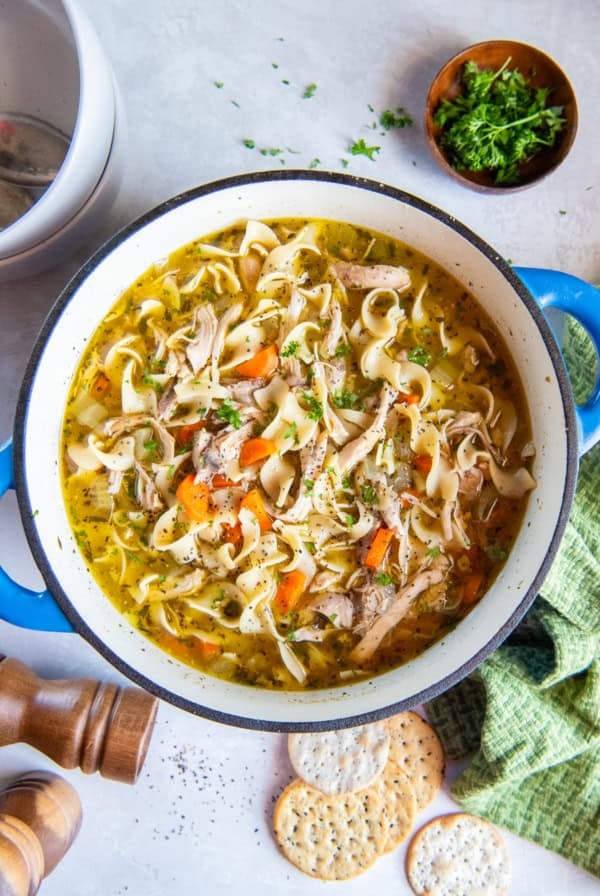 Homemade chicken noodle soup in a blue Dutch oven with egg noodles, shredded chicken, carrots, celery, and herbs, with crackers and fresh parsley nearby.