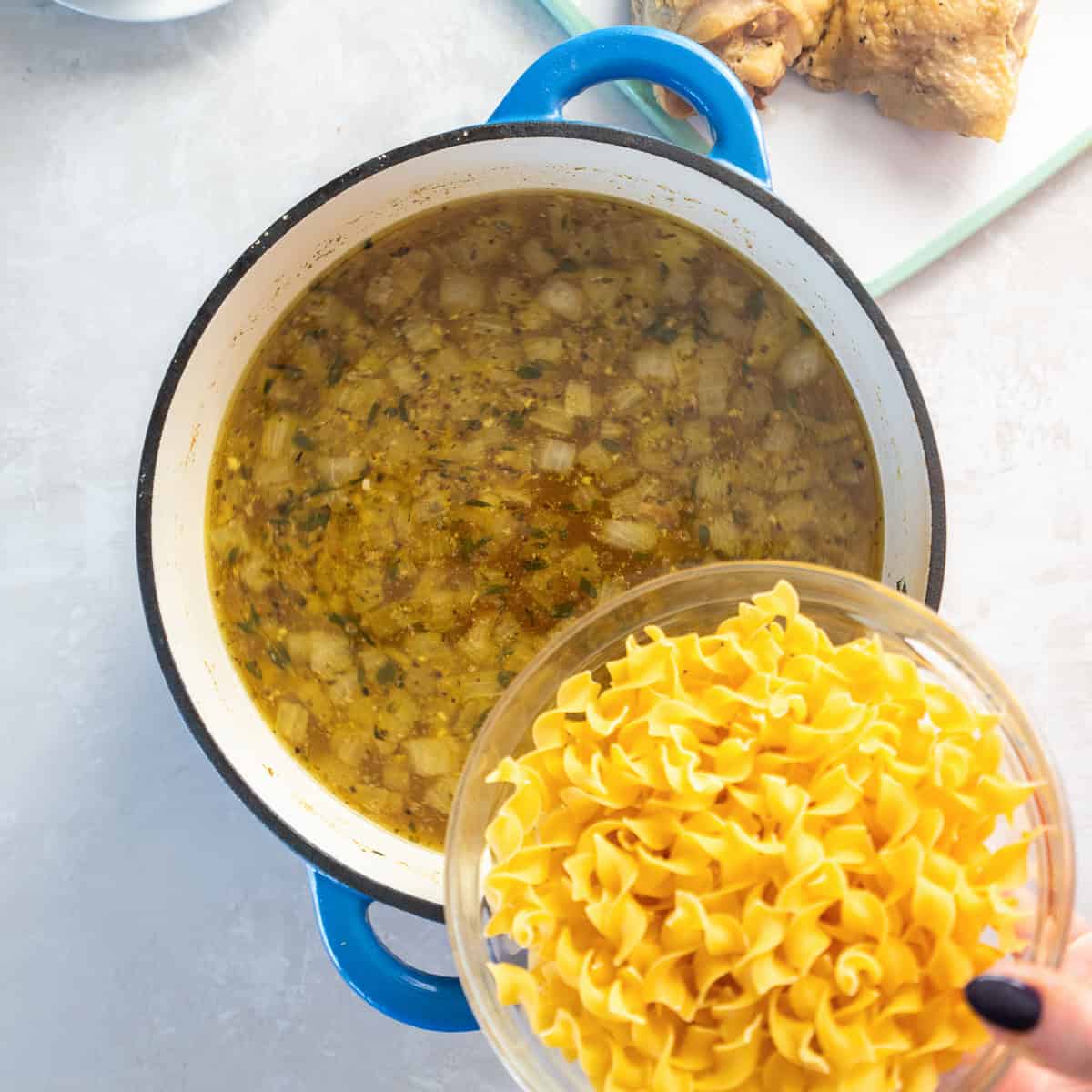 A bowl of egg noodles hovering over a pot of chicken soup broth.