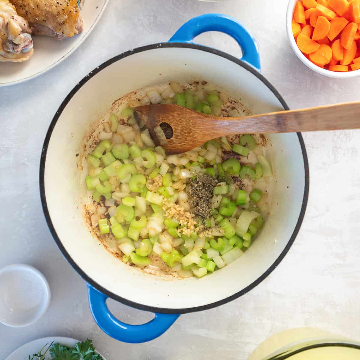 A wooden spoon stirring chopped onion and celery in a Dutch oven.