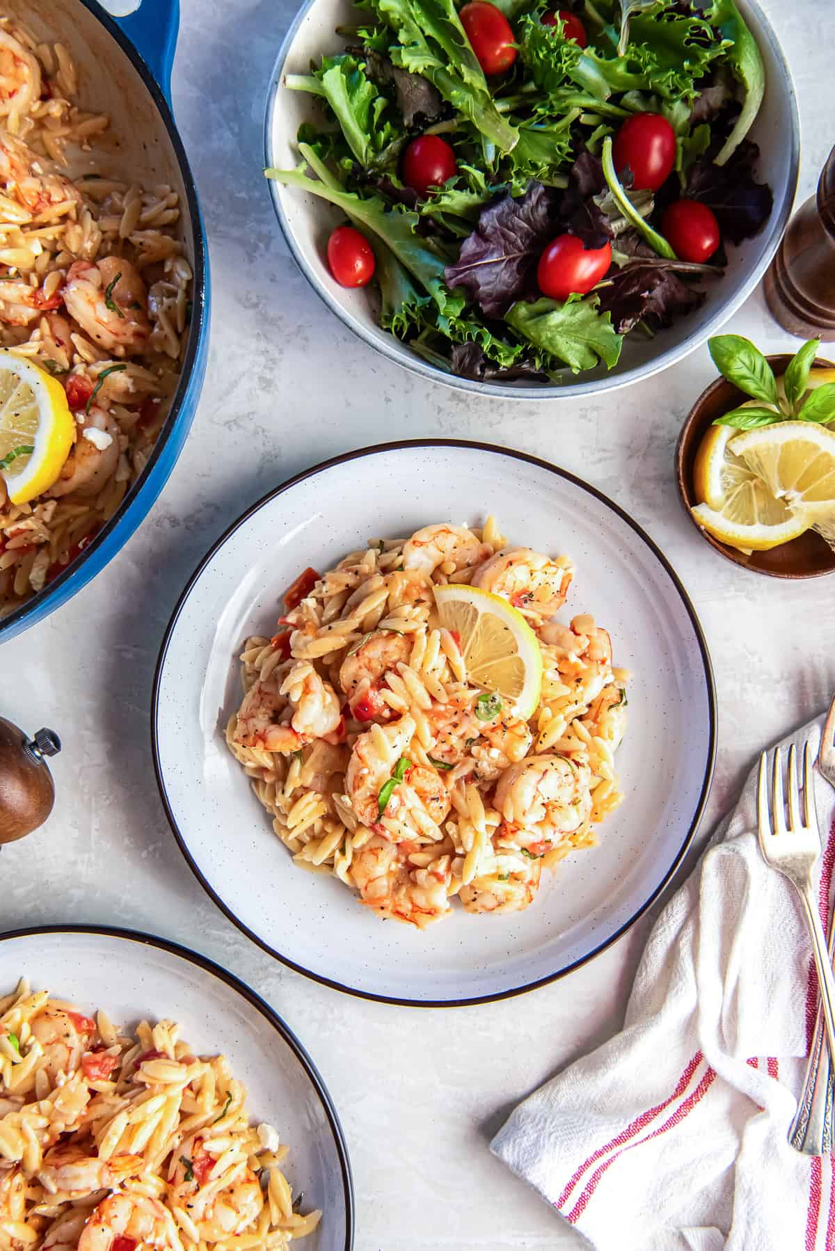 Plate of orzo pasta with shrimp, tomatoes, feta, and lemon served alongside a simple green salad, with the skillet of shrimp orzo visible in the background.