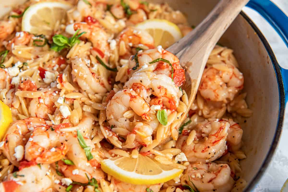 Shrimp orzo pasta with tomatoes feta and basil in a pan with a spoon lifting a serving.