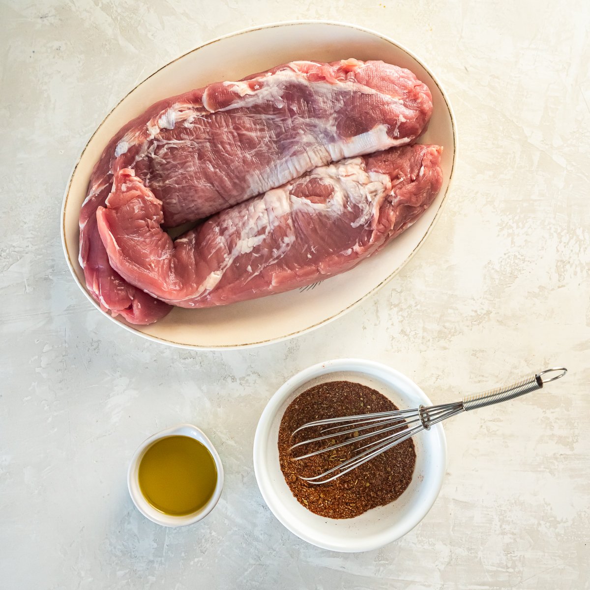 Uncooked pork tenderloins on a plate next to a small bowls of olive oil and a spice mix.