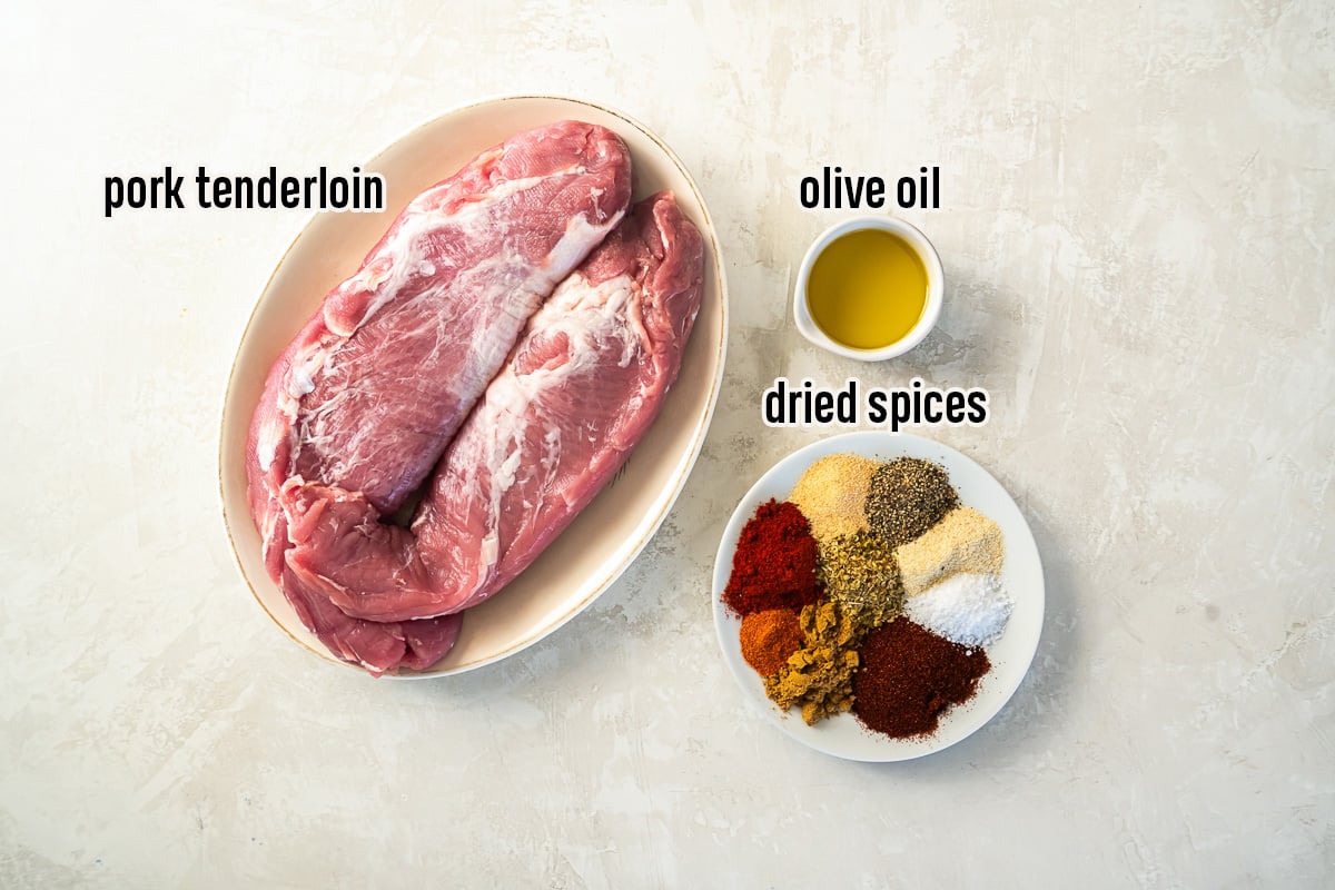 Two uncooked pork tenderloins on a plate next to a small bowl of oil and a plate full of dried spices.