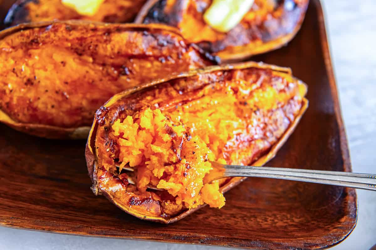Baked sweet potato halves with caramelized edges and a soft, creamy center on a wooden platter and a fork lifting a bite