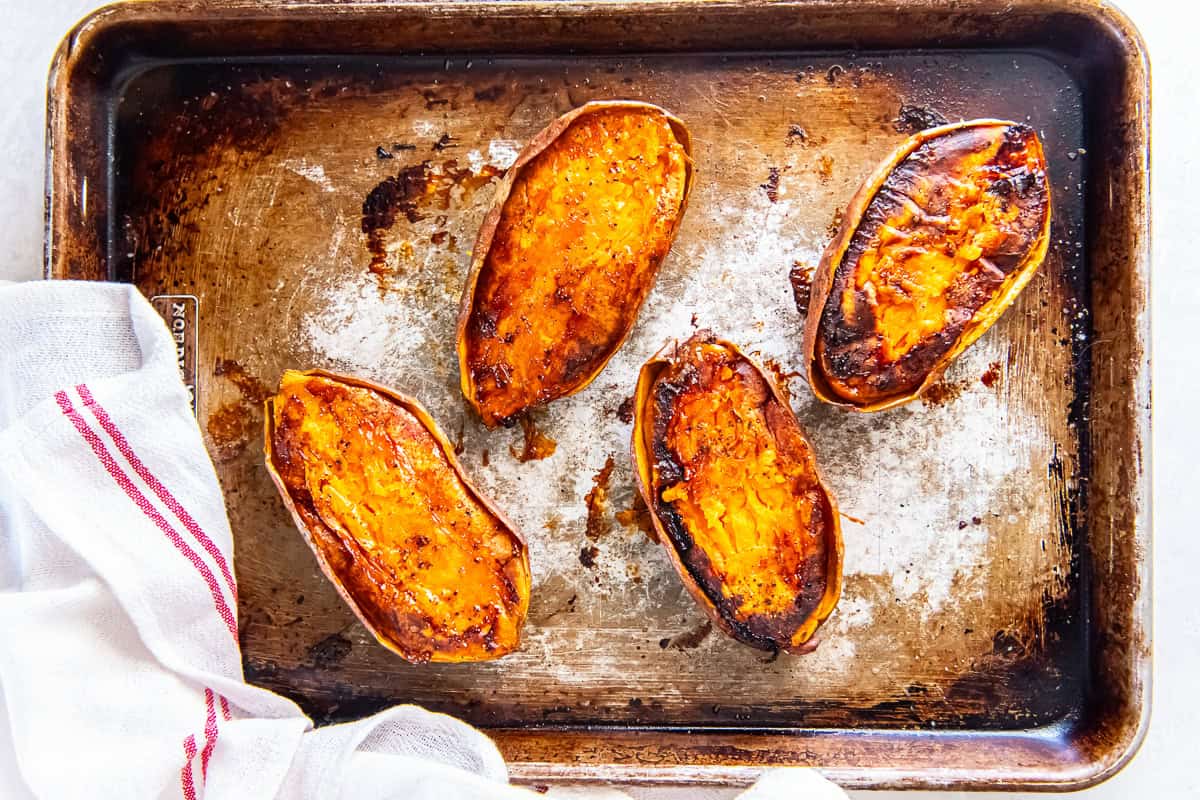 Caramelized baked sweet potato halves on a well-used sheet pan with a striped kitchen towel nearby