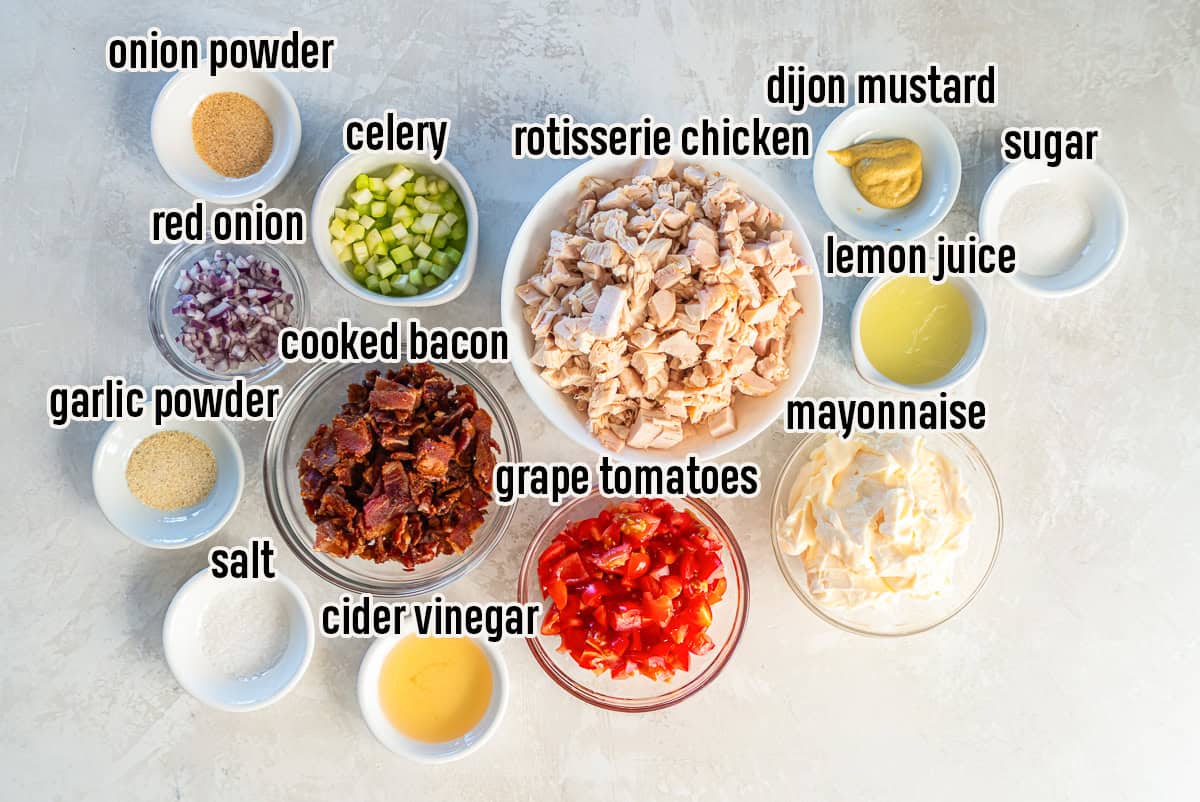 Ingredients for BLT chicken salad including chopped rotisserie chicken, cooked bacon, grape tomatoes, celery, red onion, mayonnaise, Dijon mustard, lemon juice, apple cider vinegar, sugar, garlic powder, onion powder, and salt arranged in small bowls.
