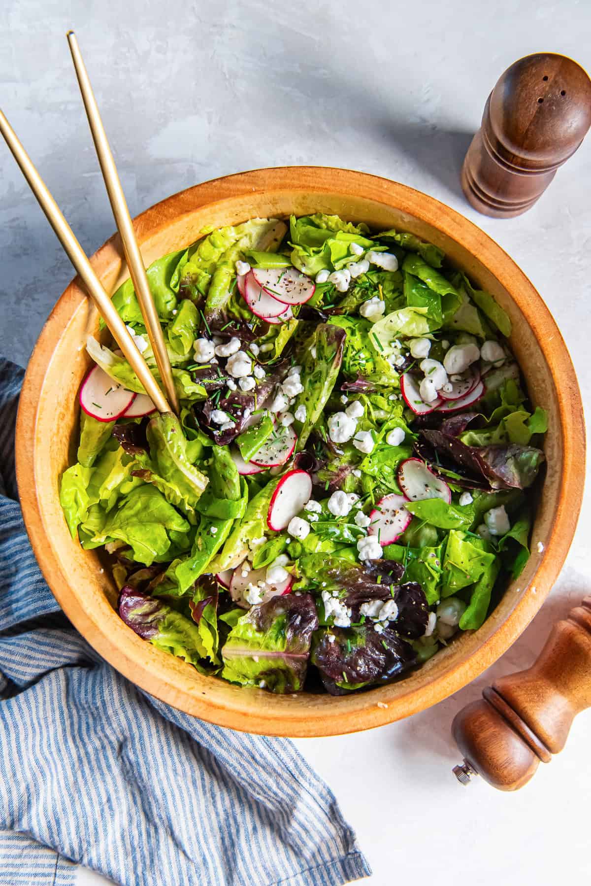 Butter lettuce salad with snap peas, radishes, goat cheese, and chives in a wooden salad bowl with salad spoons on a light countertop.