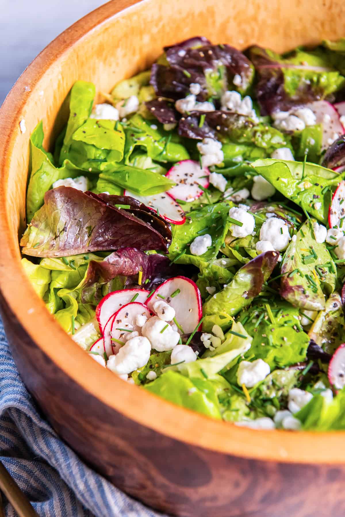 Butter lettuce salad with snap peas and radishes topped with goat cheese and chives in Champagne vinaigrette in a wood bowl.