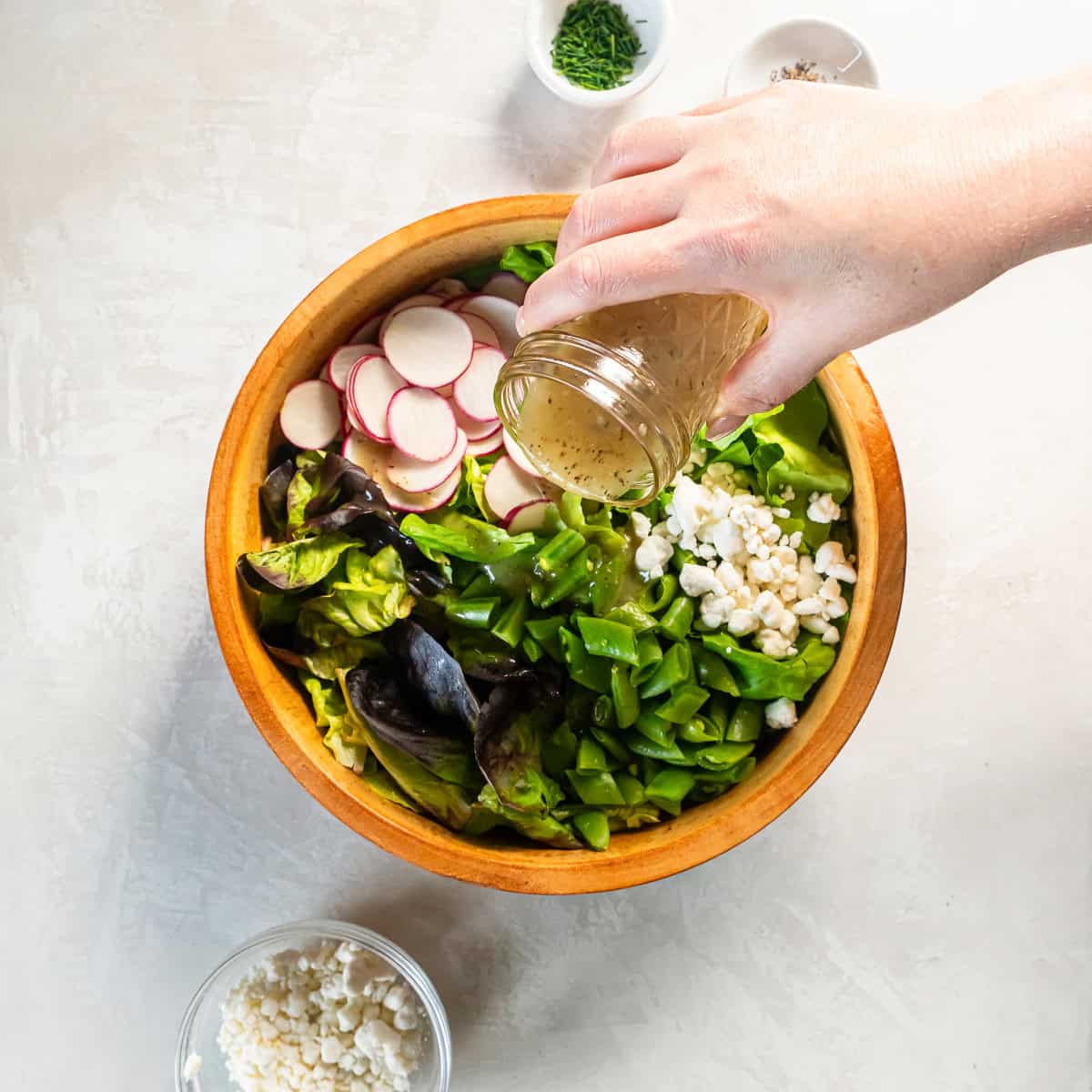 Pouring Champagne vinaigrette over butter lettuce, snap peas, radishes, and goat cheese in a wooden bowl.