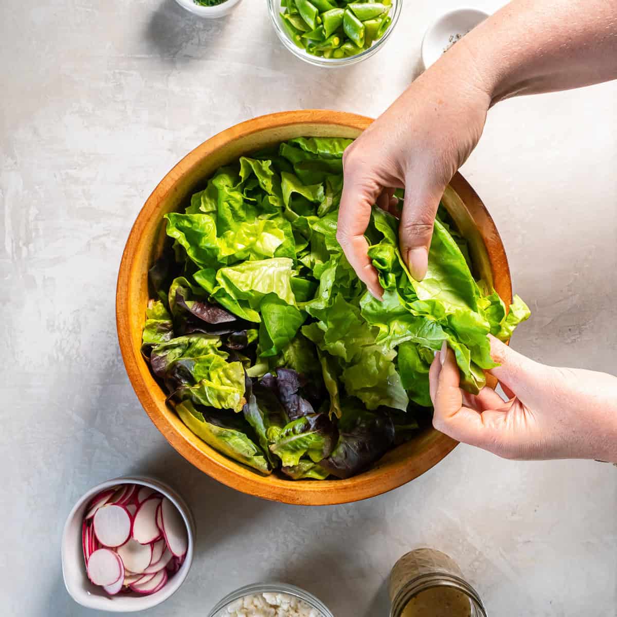 Hands tearing butter lettuce leaves into bite-size pieces in a wooden bowl with baby lettuce blend for butter lettuce salad.