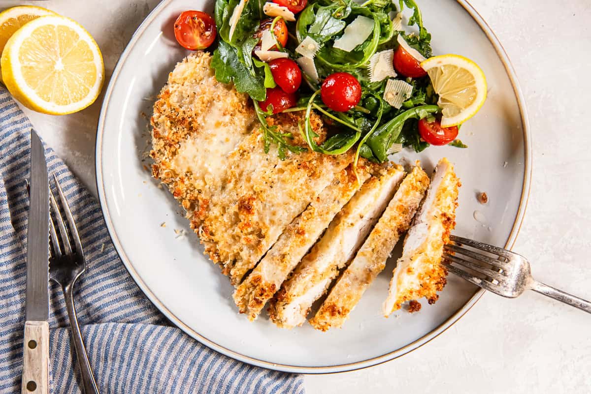 Sliced Chicken Milanese with crispy Parmesan breadcrumb coating served with arugula salad, cherry tomatoes, and lemon wedges.