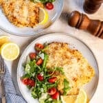 Two plates of Chicken Milanese with arugula salad, cherry tomatoes, shaved Parmesan, and lemon wedges.
