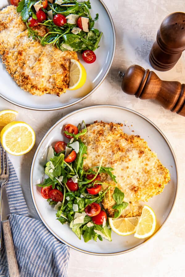 Two plates of Chicken Milanese with arugula salad, cherry tomatoes, shaved Parmesan, and lemon wedges.