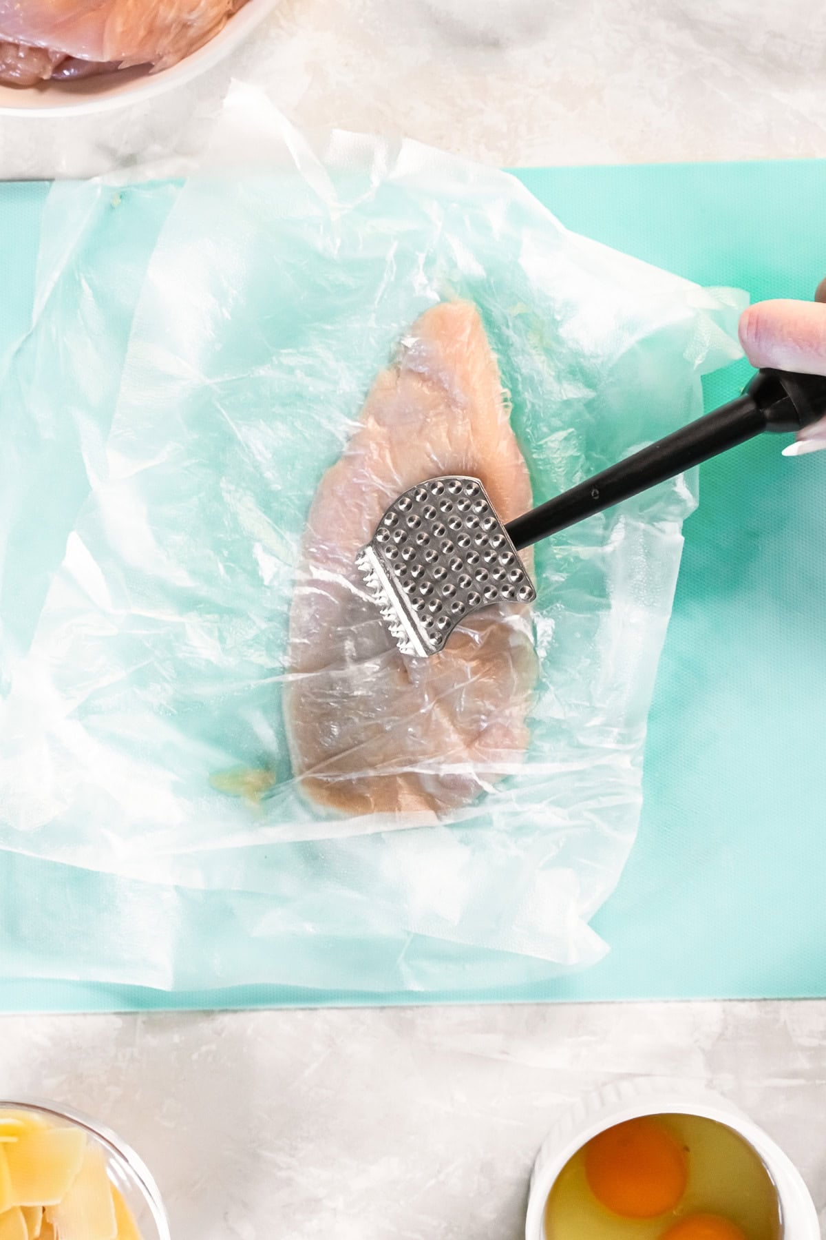 Chicken breast being pounded thin with a meat mallet between sheets of plastic wrap.