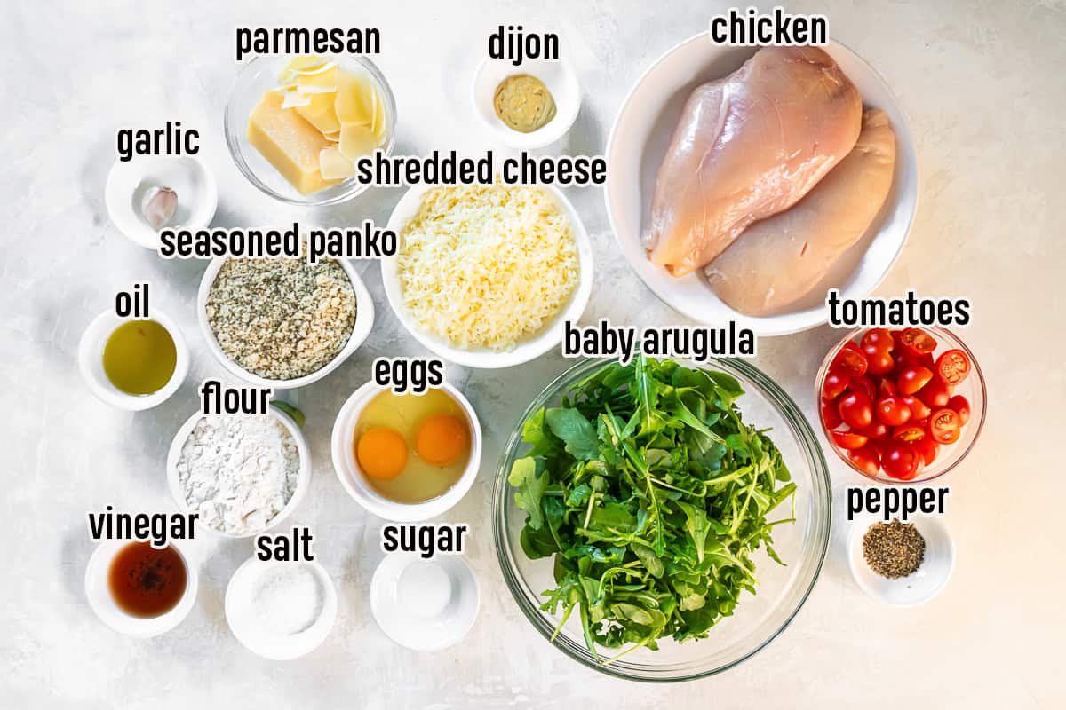 Ingredients for Chicken Milanese including chicken breasts, Italian seasoned panko, flour, eggs, Parmesan, shredded cheese, Dijon mustard, baby arugula, cherry tomatoes, olive oil, red wine vinegar, garlic, salt, pepper, and sugar arranged on a countertop.