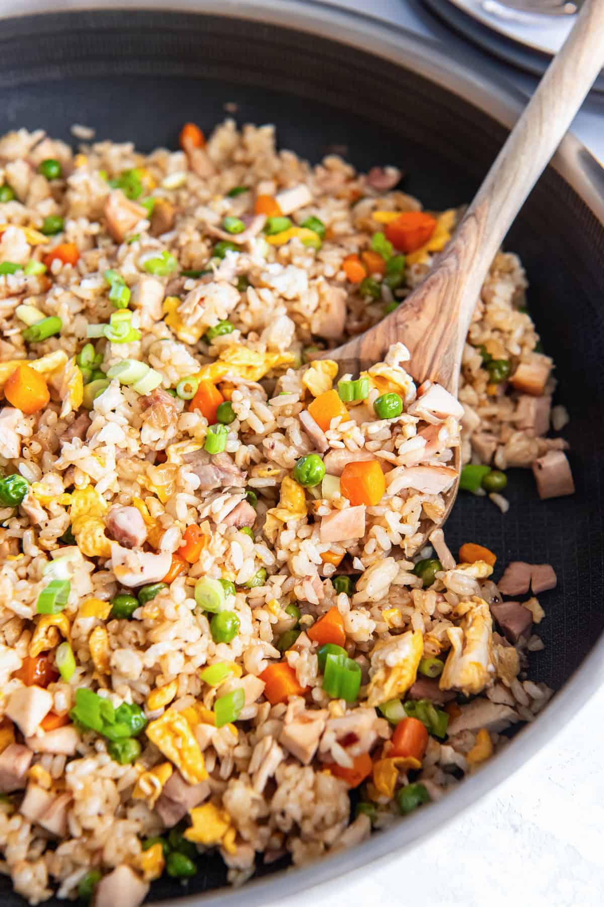 Chicken fried rice cooking in a skillet with a wooden spoon, peas, carrots, scrambled eggs, and chopped chicken.