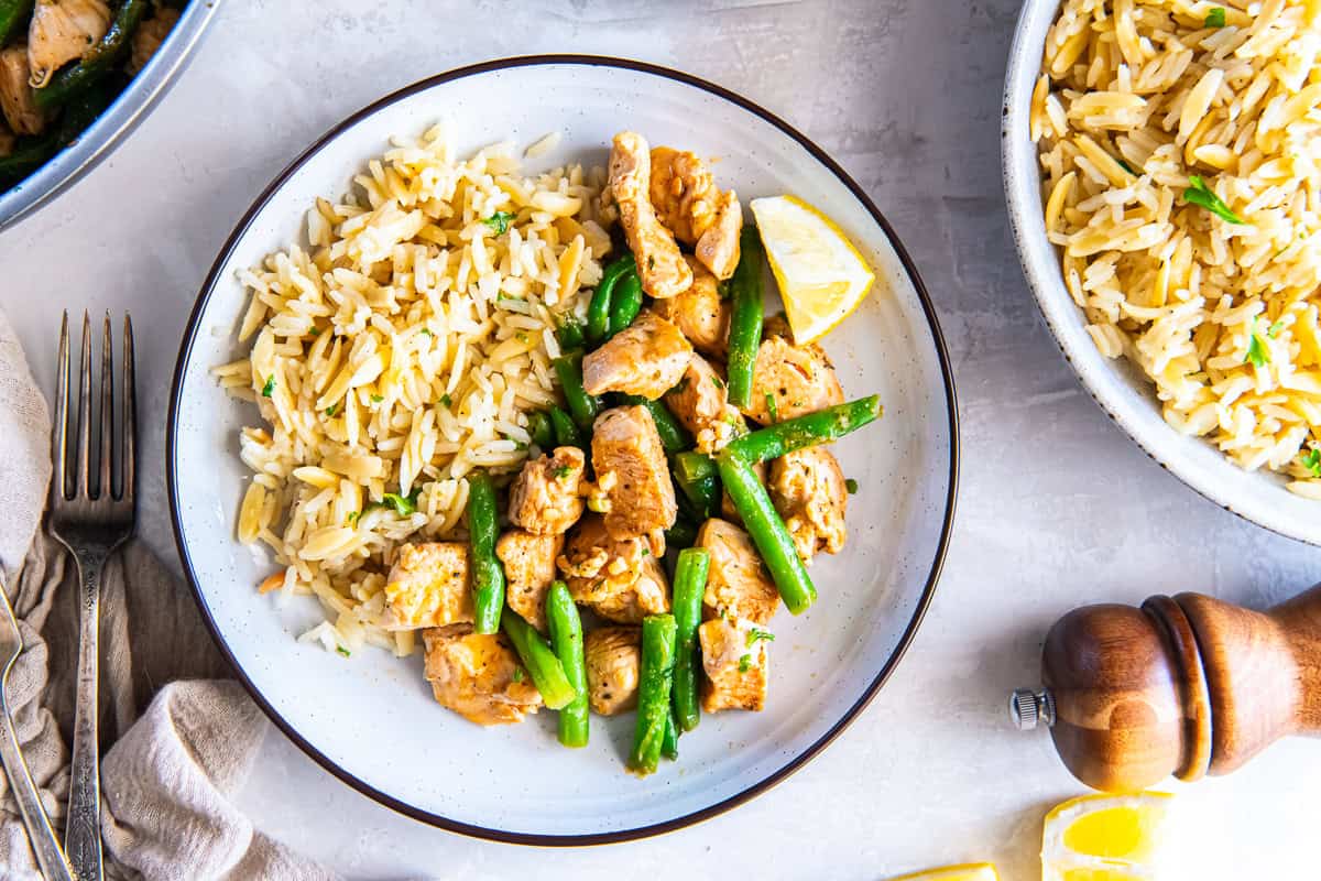 Garlic butter chicken and green beans served with rice on a plate, garnished with a lemon wedge, with a bowl of rice and pepper mill in the background.