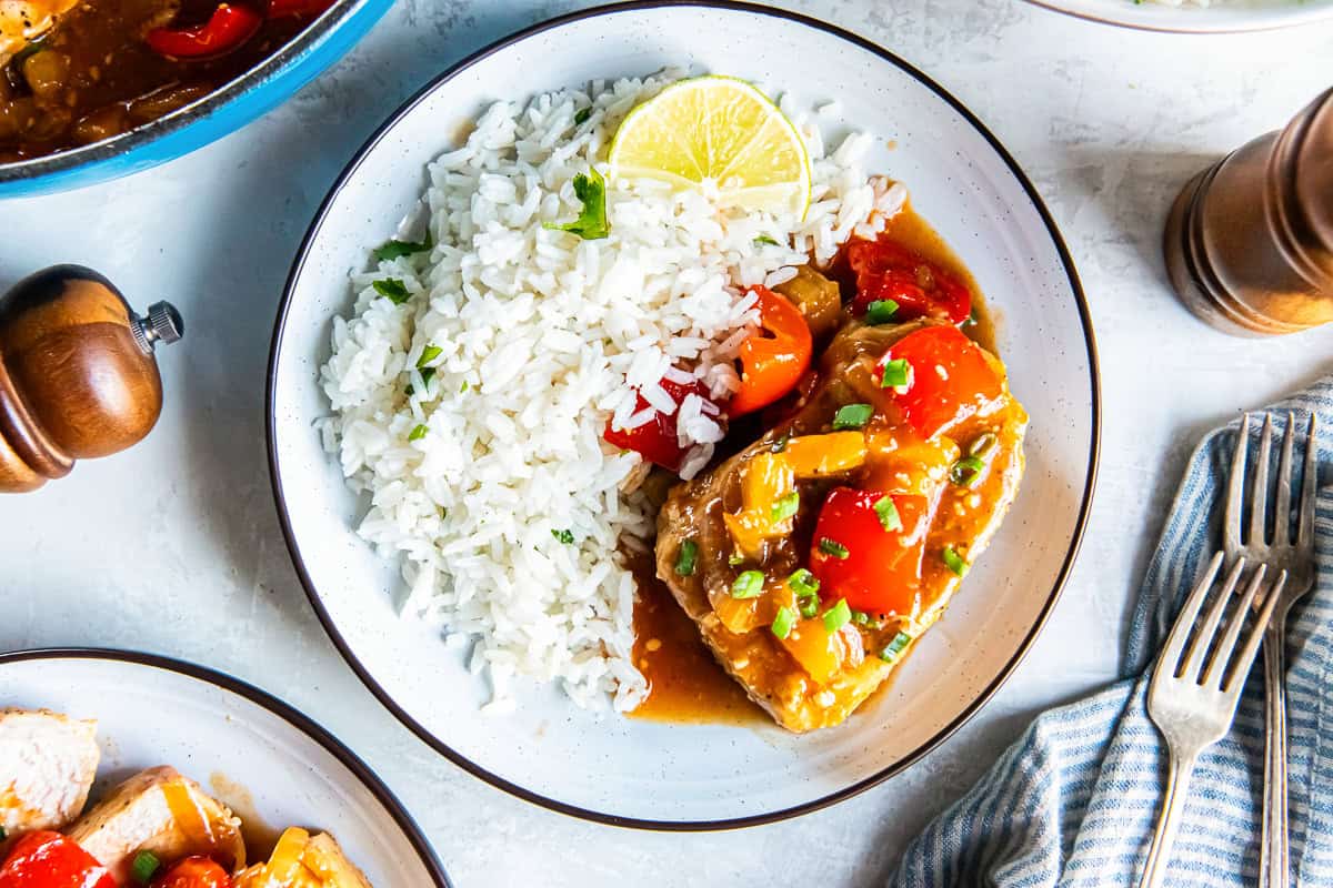 Hawaiian pork chop topped with pineapple tidbits, red bell pepper, and onion in a glossy sauce, served with white rice and a lime wedge on a plate.