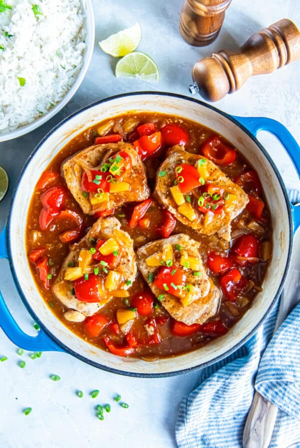 Skillet of Hawaiian pork chops in a glossy pineapple sauce with red bell pepper, onion, and pineapple tidbits, served with a bowl of white rice and lime wedges on the side.