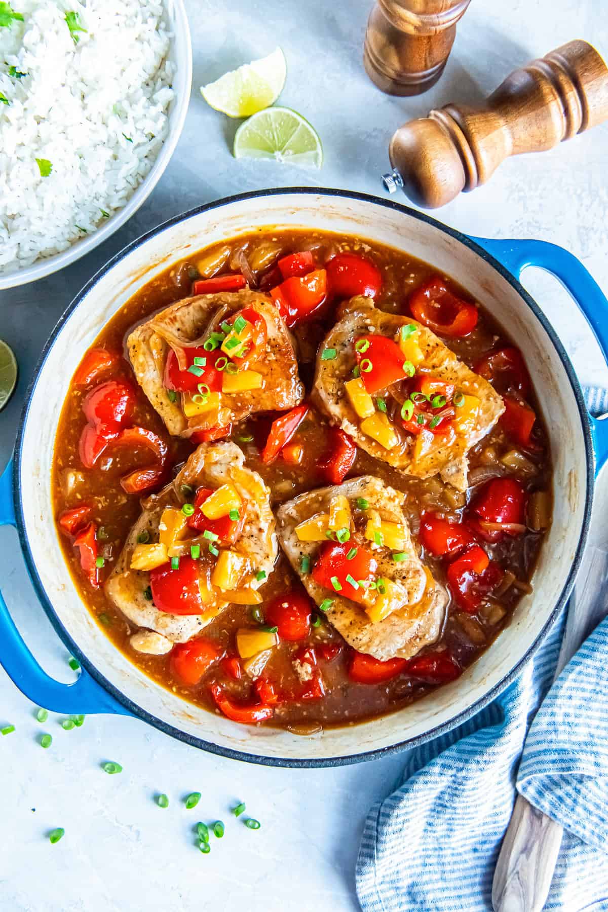 Skillet of Hawaiian pork chops in a glossy pineapple sauce with red bell pepper, onion, and pineapple tidbits, served with a bowl of white rice and lime wedges on the side.