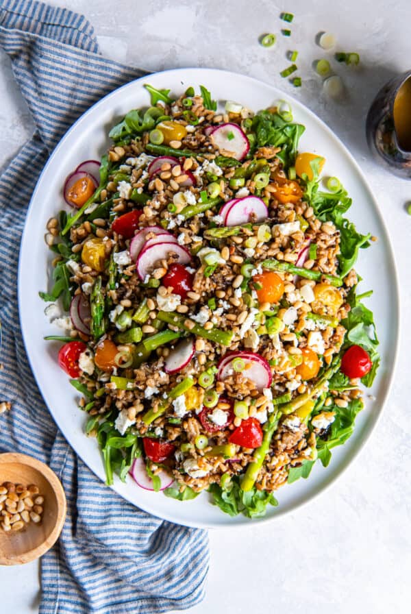 Spring grain salad with asparagus, radishes, cherry tomatoes, goat cheese, pine nuts, and arugula tossed in apple cider vinaigrette on a serving platter.