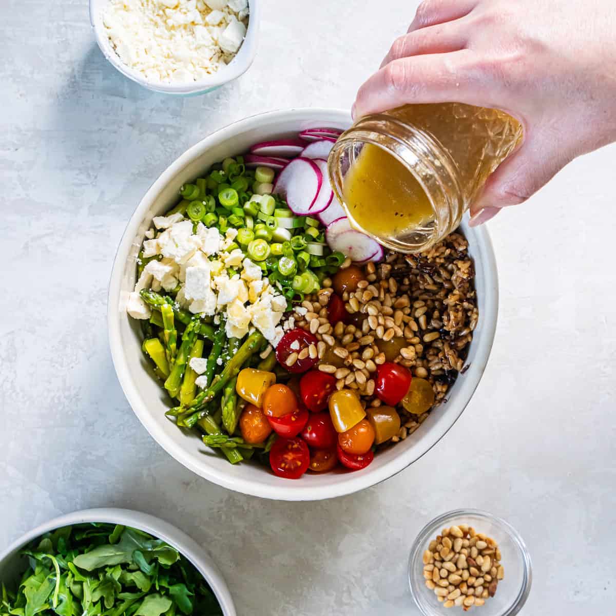 Pouring apple cider vinaigrette over mixed grains, asparagus, tomatoes, radishes, green onions, and goat cheese.