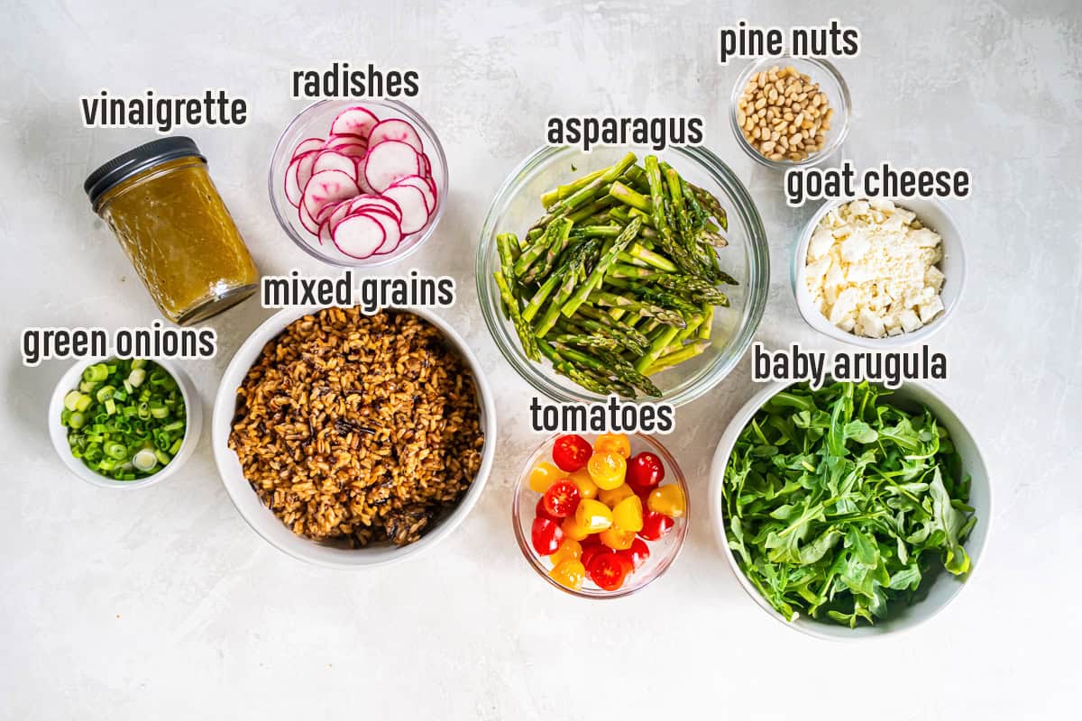 Spring Grain Salad ingredients including mixed grains, asparagus, radishes, cherry tomatoes, baby arugula, green onions, pine nuts, goat cheese, and apple cider vinaigrette arranged in bowls on a light background.