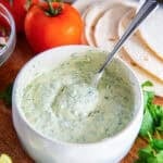 Creamy cilantro lime sauce in a bowl with a spoon, served with tortillas, tomatoes, lime wedges, and fresh cilantro