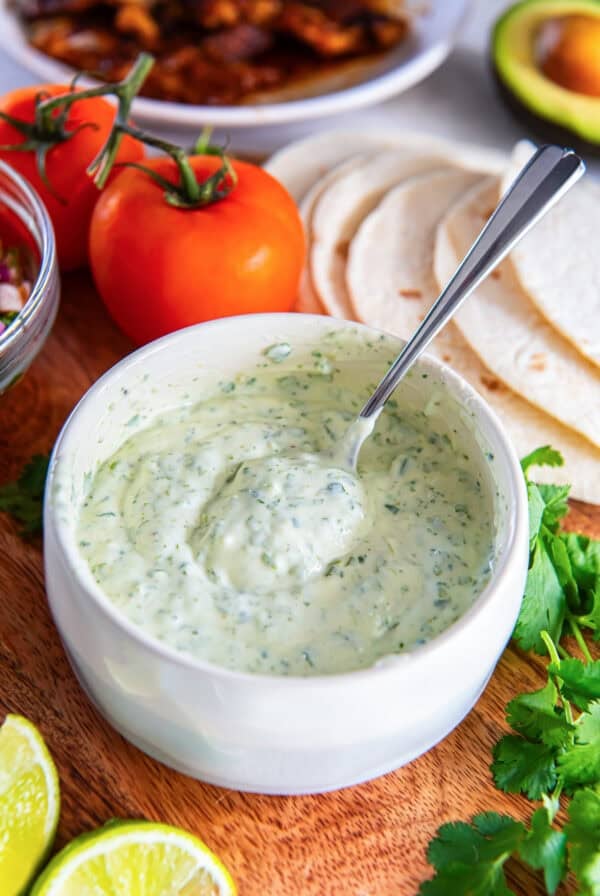 Creamy cilantro lime sauce in a bowl with a spoon, served with tortillas, tomatoes, lime wedges, and fresh cilantro