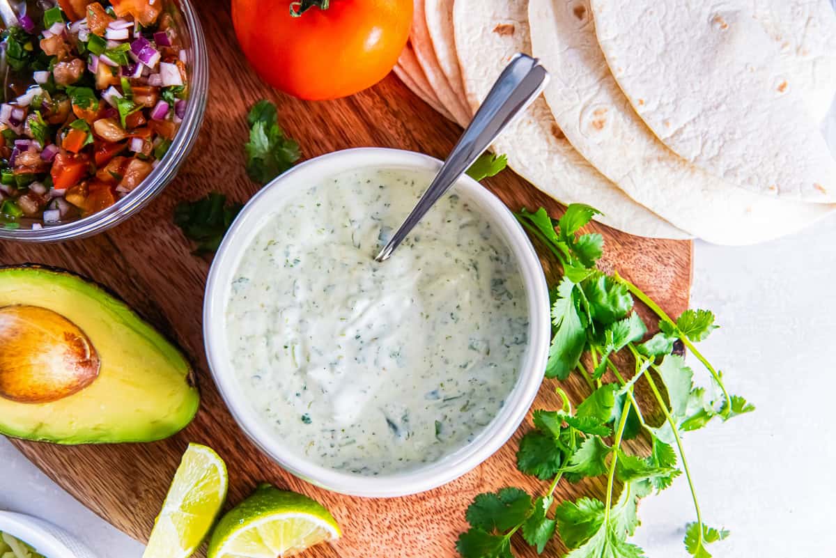 Cilantro lime sauce in a bowl with tortillas, pico de gallo, avocado, lime wedges, and fresh cilantro on a wooden board.