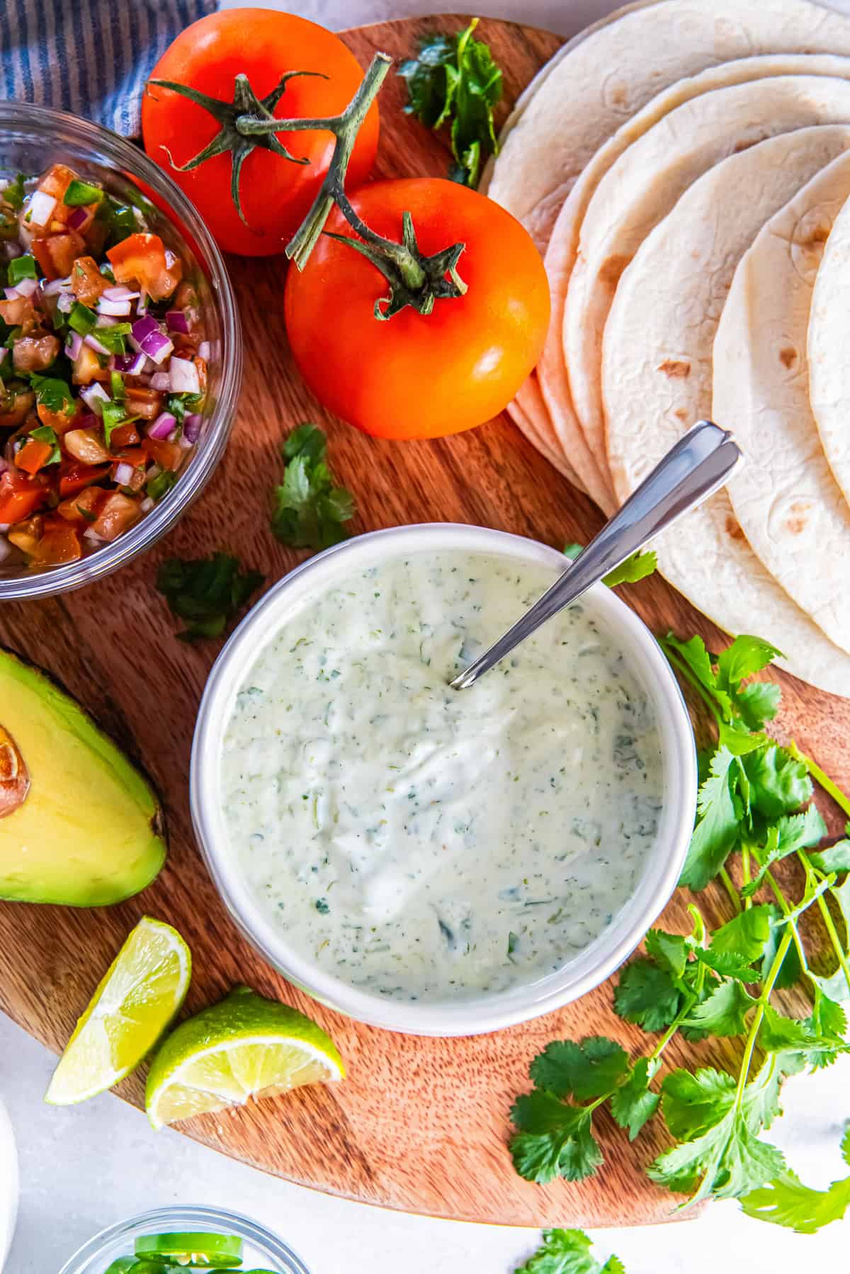 Cilantro lime sauce in a bowl with tortillas, pico de gallo, avocado, lime wedges, and fresh cilantro on a wooden board.