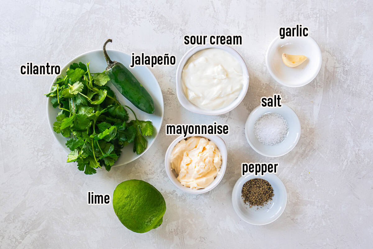 Cilantro lime sauce ingredients including cilantro, jalapeño, lime, sour cream, mayonnaise, garlic, salt, and pepper arranged on a white surface