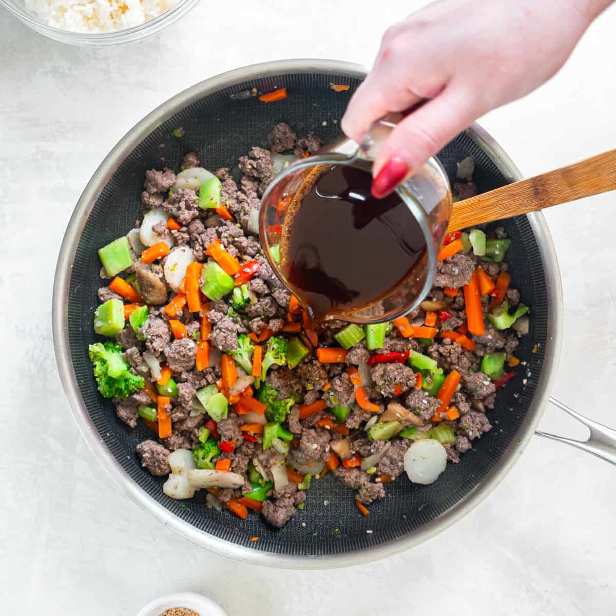 Pouring teriyaki sauce over cooked ground beef and vegetables in skillet.
