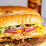 Italian chicken sub sandwich on a soft sandwich roll with melted provolone, lettuce, tomato, red onion, and banana peppers, with iced tea in the background.