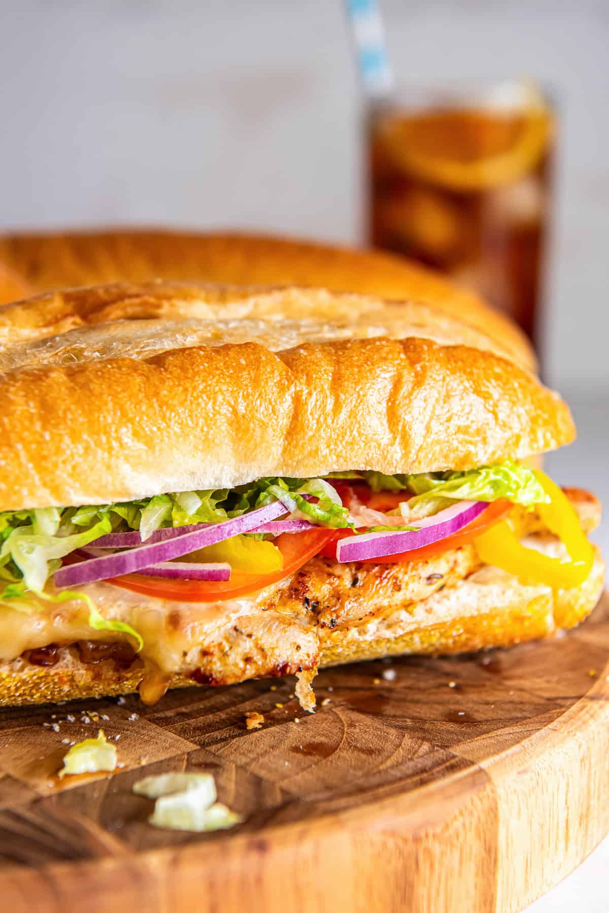 Italian chicken sub sandwich on a soft sandwich roll with melted provolone, lettuce, tomato, red onion, and banana peppers, with iced tea in the background.