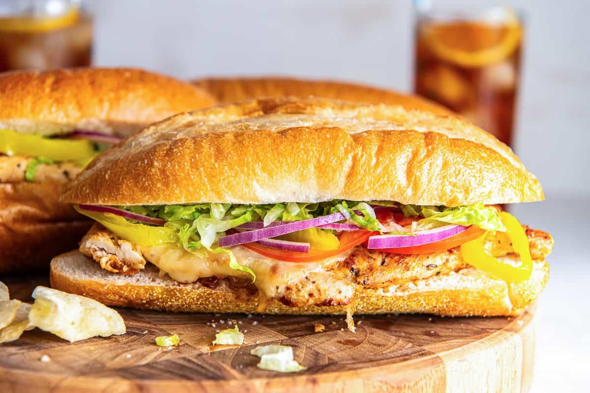 Italian chicken sub sandwich on a soft sandwich roll with melted provolone, lettuce, tomato, red onion, and banana peppers, served with chips.