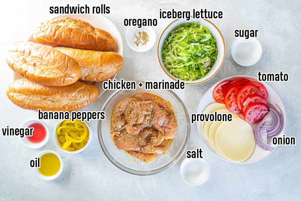 Ingredients for Italian chicken subs including sandwich rolls, marinated chicken, iceberg lettuce, tomato, onion, provolone, banana peppers, oil, vinegar, oregano, sugar, and salt.