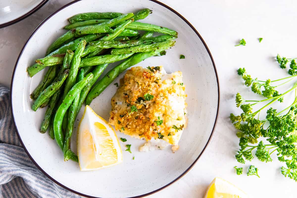 Parmesan crusted fish served with green beans and lemon wedges on a plate.