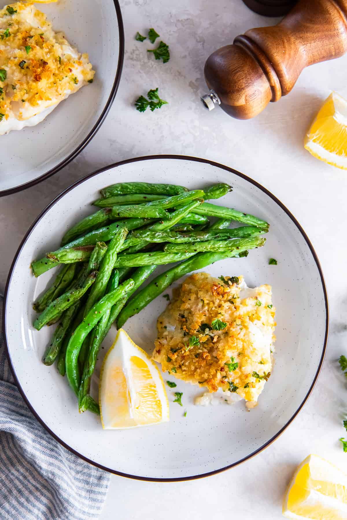 Plate of Parmesan crusted fish with green beans and lemon wedges, with a pepper mill in the background.