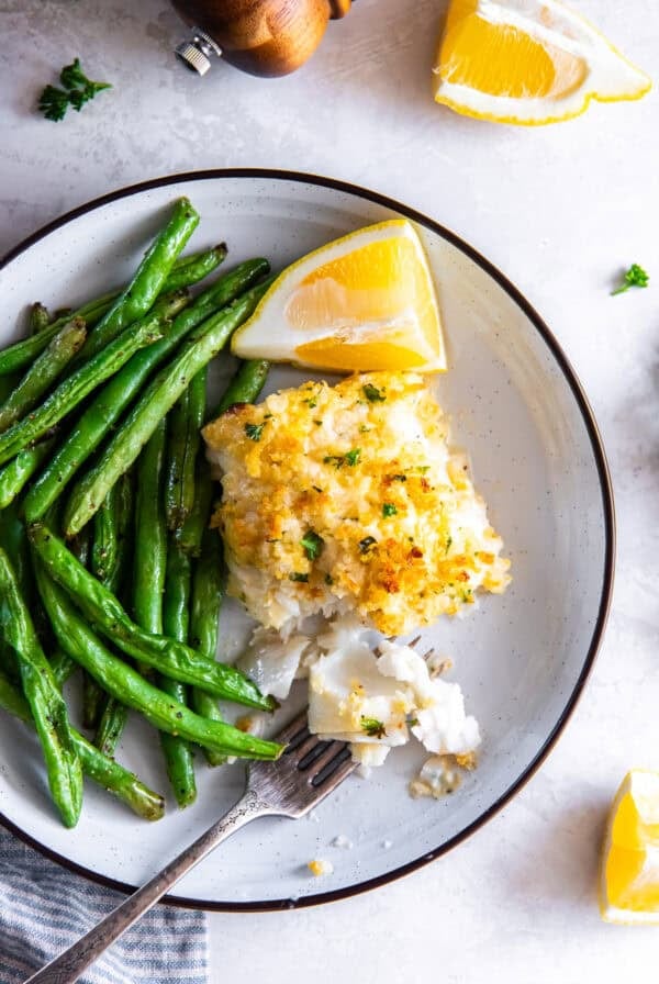 Flaky Parmesan crusted fish served with green beans and lemon wedges, with a fork breaking into the fish.