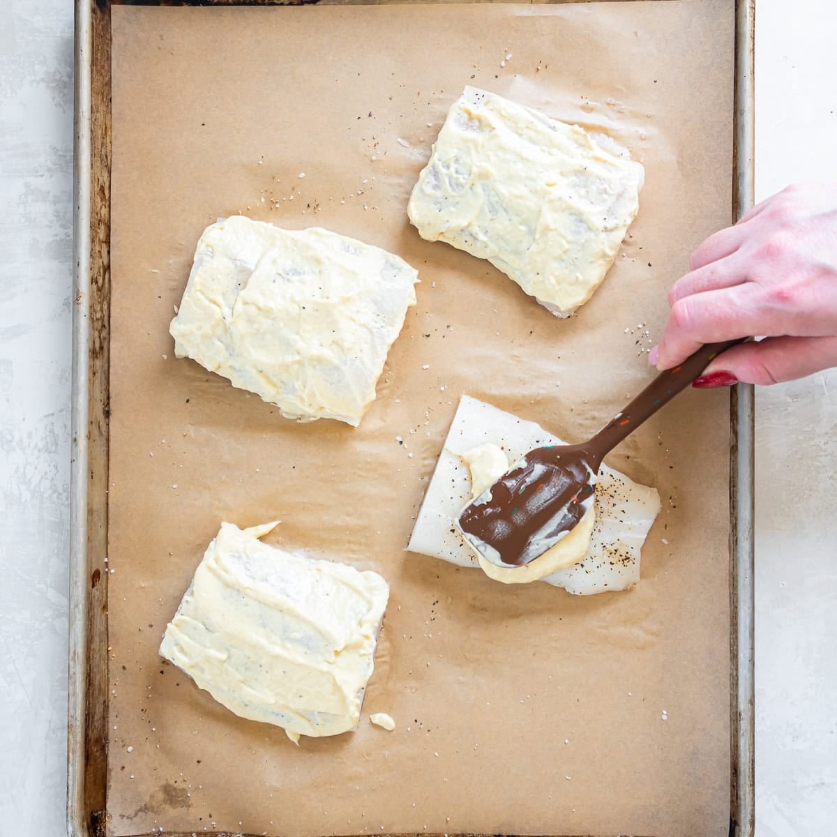 Spreading the creamy Dijon mixture over seasoned fish fillets on a baking sheet.