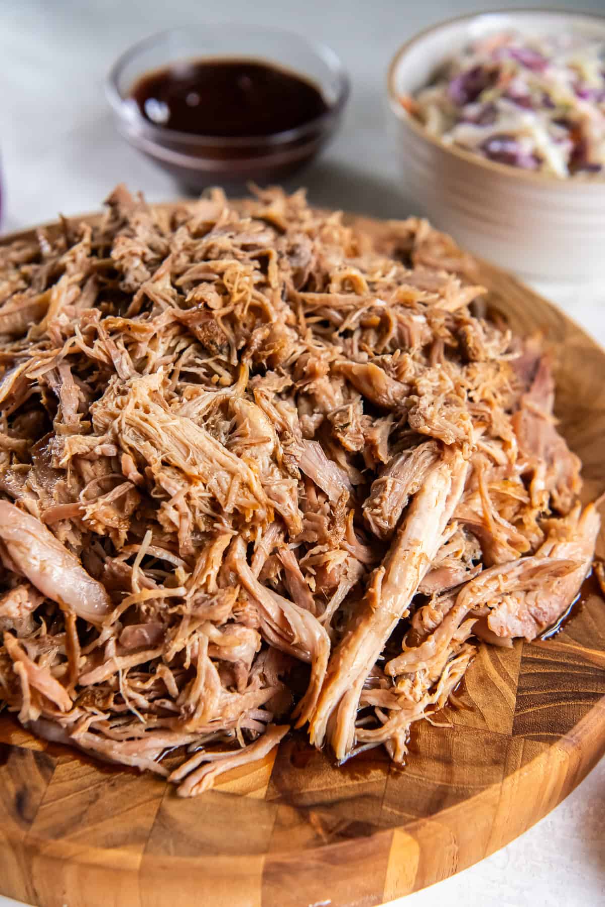 Slow cooker pulled pork served on a wooden board with barbecue sauce and coleslaw in the background.