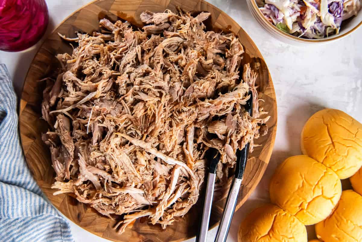 Shredded slow cooker pulled pork on a wooden board with slider buns and coleslaw on the side.