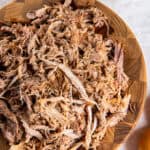 Slow cooker pulled pork shredded and piled on a wooden board with barbecue sauce and coleslaw in the background.