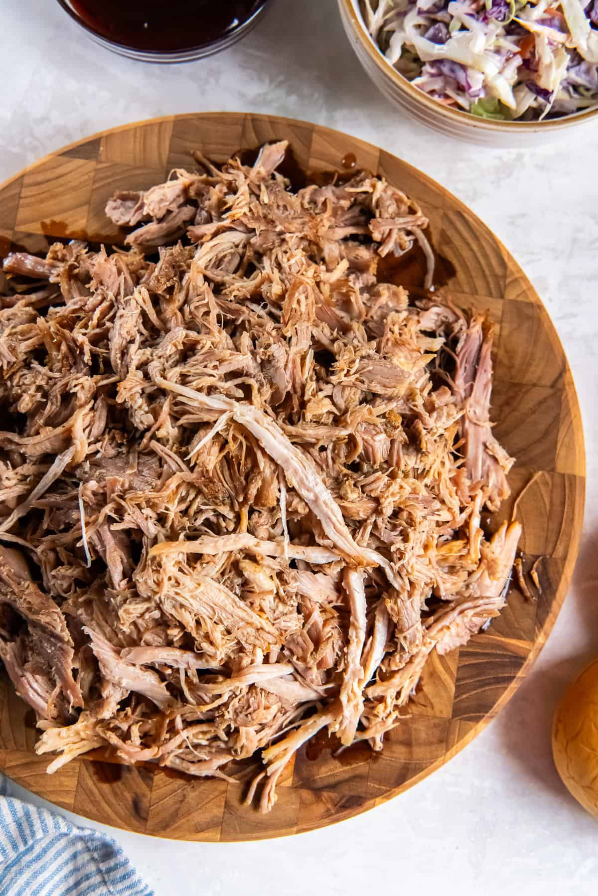 Slow cooker pulled pork shredded and piled on a wooden board with barbecue sauce and coleslaw in the background.