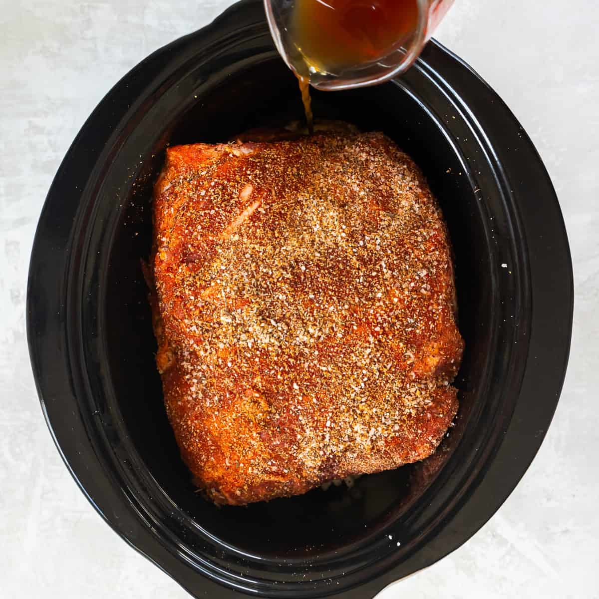 Seasoned pork shoulder placed over onions in a slow cooker with cooking liquid being added.