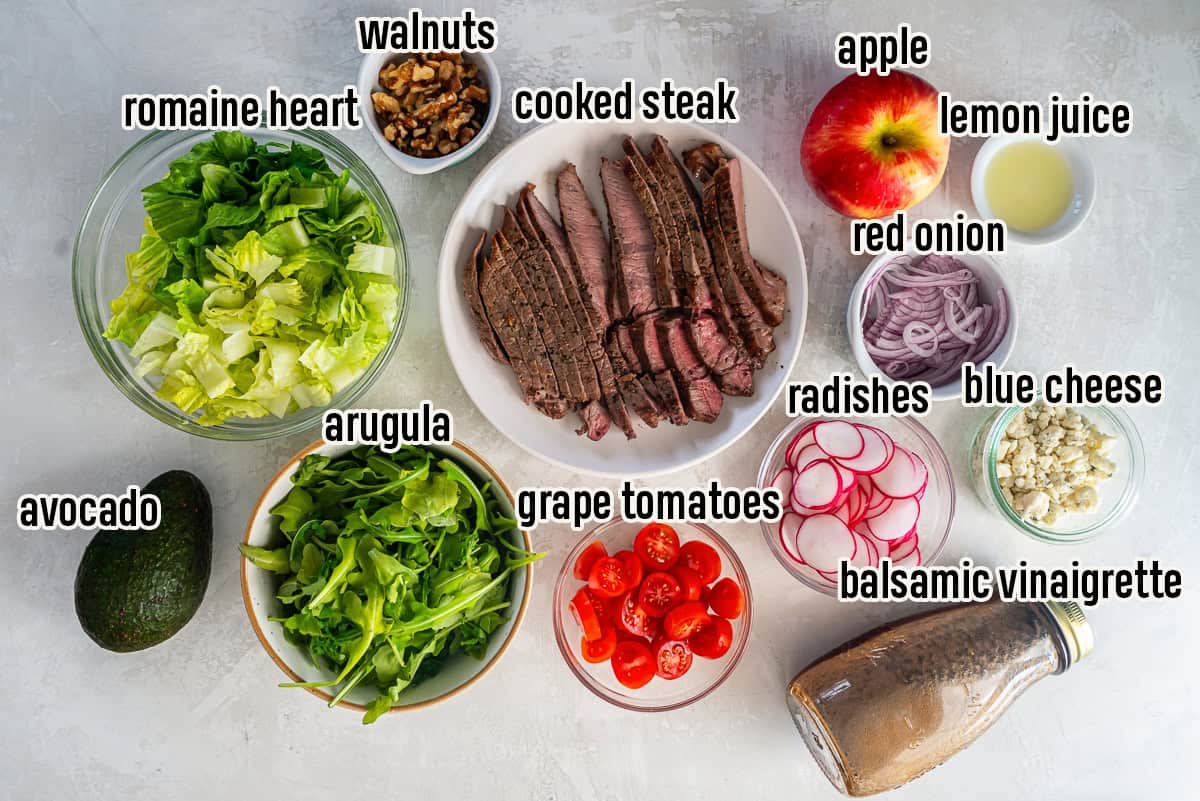 Steak salad ingredients including romaine, arugula, cooked steak, apple, avocado, tomato, red onion, radish, blue cheese, walnuts, and balsamic vinaigrette.